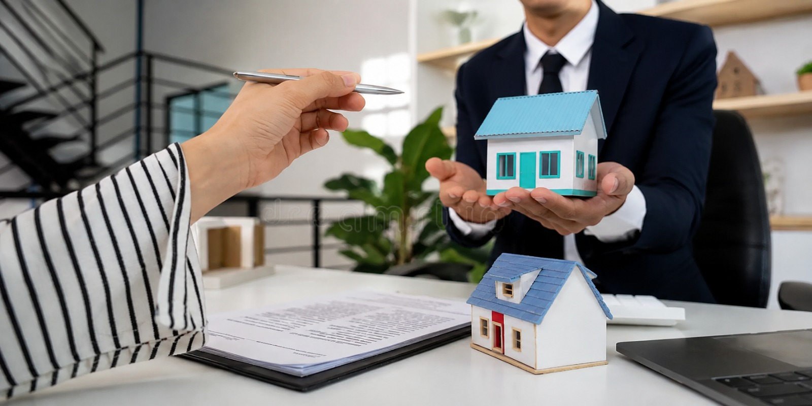 Property agent in a suit holding a model house across a desk from a client signing documents